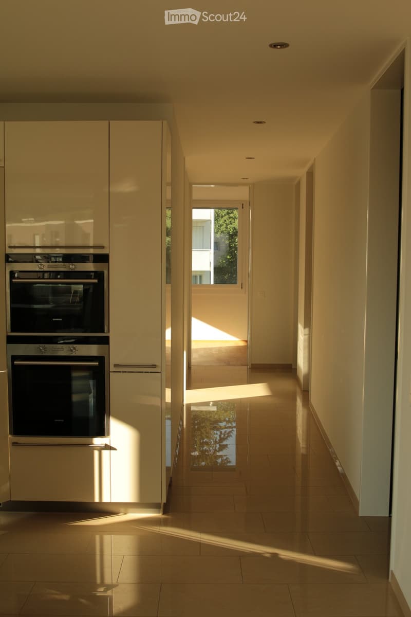 Modern hallway with light-colored walls, tiled floor, and built-in cabinets with stainless steel appliances visible on the left side. Sunlight streams in from an open doorway at the end of the corridor.