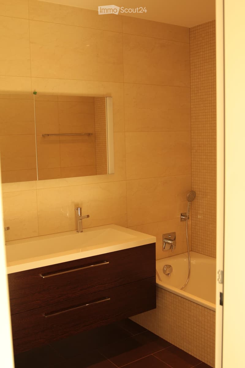 Modern bathroom with a dark wooden vanity, white sink, chrome faucet, bathtub, and showerhead. Light-colored tiles and a large mirror complete the space.