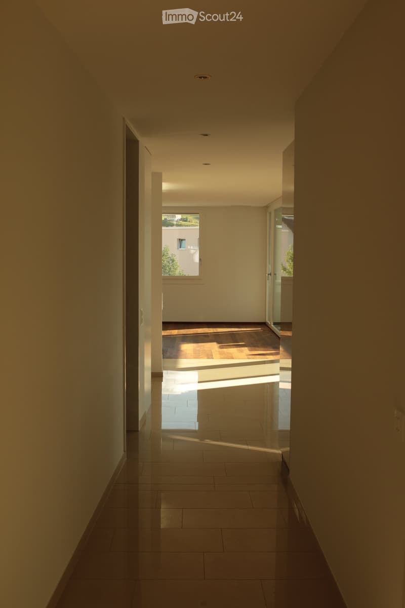 Modern hallway with tiled floor, recessed lighting, and a window at the end, leading to another room with wooden flooring.