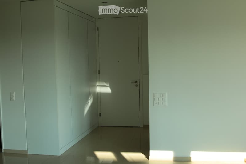 A clean, modern hallway with light green walls, tiled floor, and a closed door at the end. The space is well-lit with natural light casting shadows on the floor.