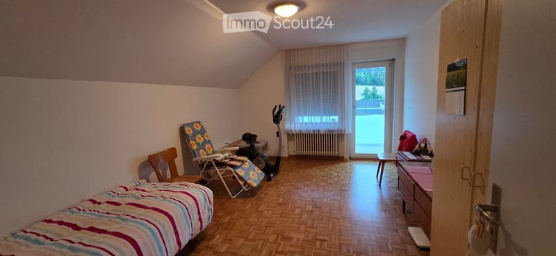 A small, cozy bedroom with wooden flooring, a bed with striped bedding, a wooden chair, a small table, an exercise bike, and a drawer unit.