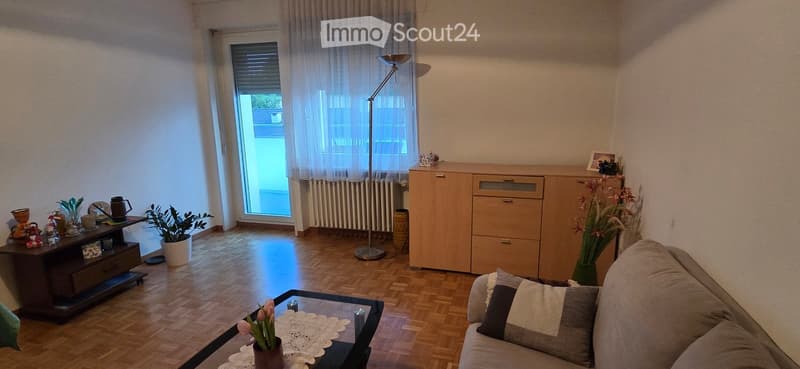 Living room with wooden flooring, a sofa with pillows, a coffee table with a lace doily and flowers, a side table with decorative items, a floor lamp, a cabinet, and potted plants.