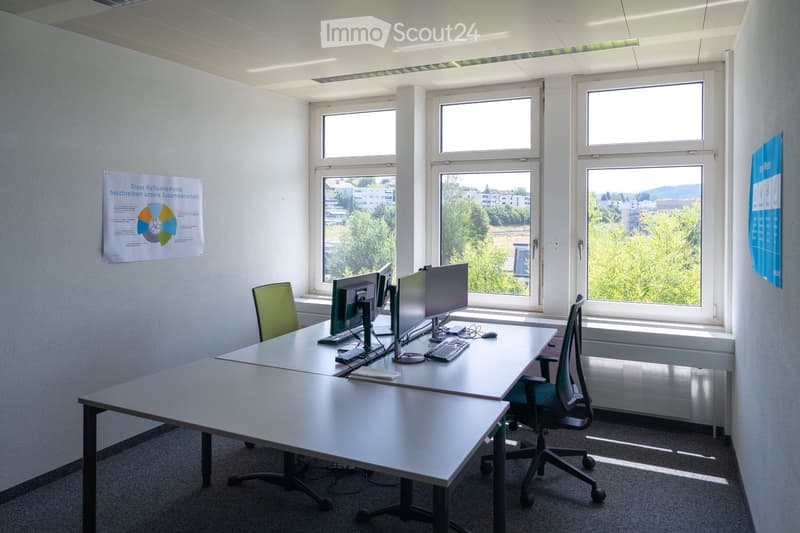 Bright office space with large windows, two desks, two chairs, two computer monitors, a keyboard, and a mouse. Posters on the walls.