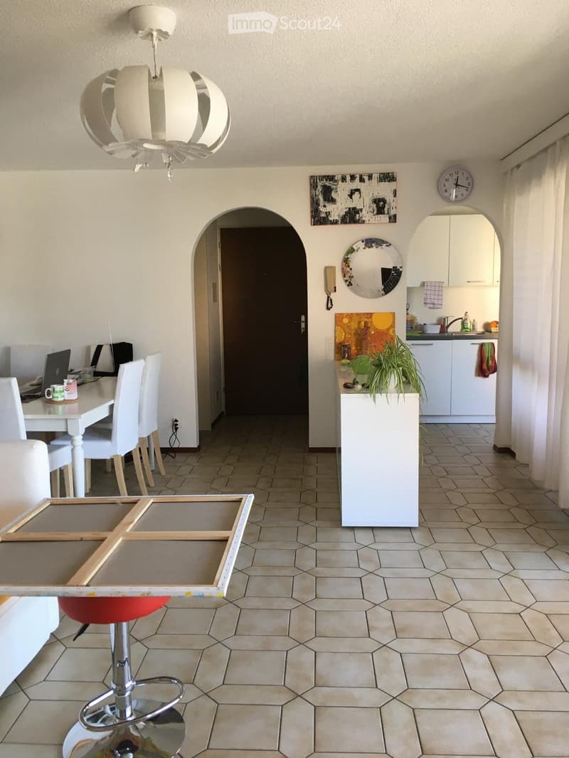 Open-plan living and dining area with a white sofa, red stool, glass coffee table, dining table with chairs, laptop, and a small kitchen area with a sink, faucet, and plant. The space features a modern chandelier and a decorative mirror.