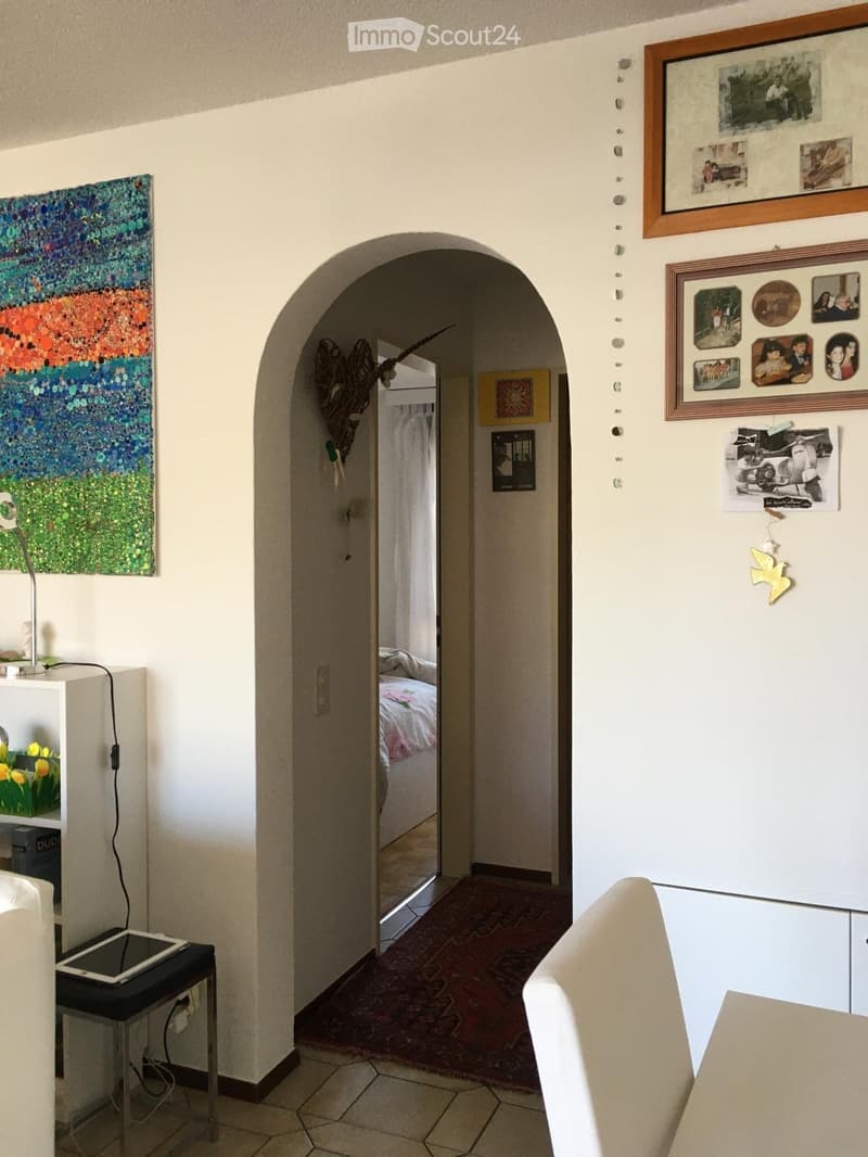 A hallway with a colorful abstract painting on the left wall, a framed collage of photos on the right, and an arched doorway leading to a bedroom with a bed and colorful bedding.