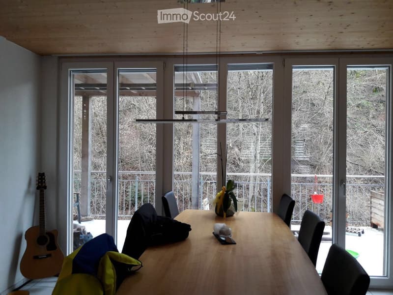 Bright dining area with large windows and sliding doors leading to a balcony, featuring a wooden dining table, chairs, a guitar, and a potted plant.