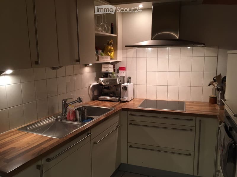 Modern kitchen with wooden countertops, stainless steel sink, coffee machine, and built-in oven. Light gray cabinets and tiled backsplash.