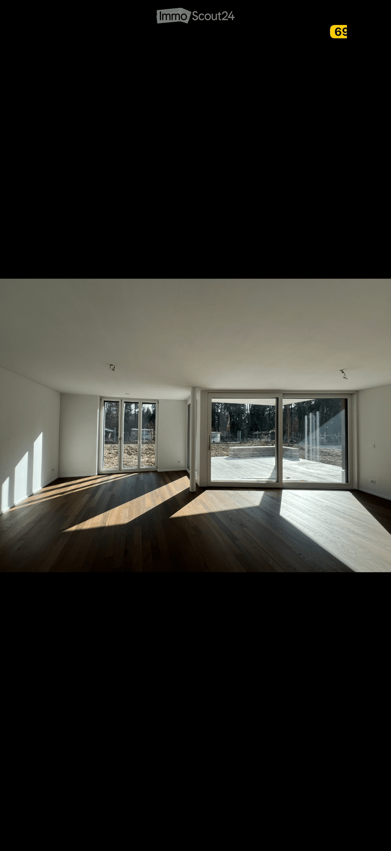 Bright, empty room with wooden flooring and large sliding glass doors leading to an outdoor deck. Sunlight streams in, casting shadows on the floor.