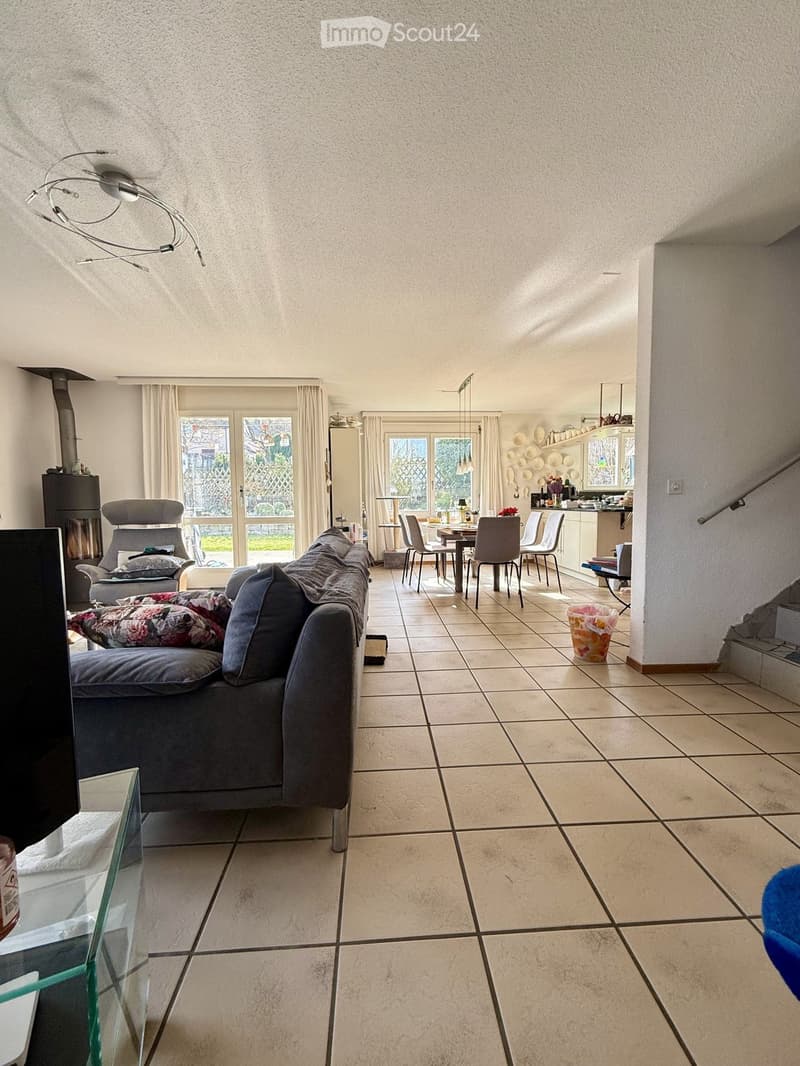 Open plan living room with dining area, fireplace, large windows, tile flooring, couch, armchair, dining table, chairs, kitchen, staircase