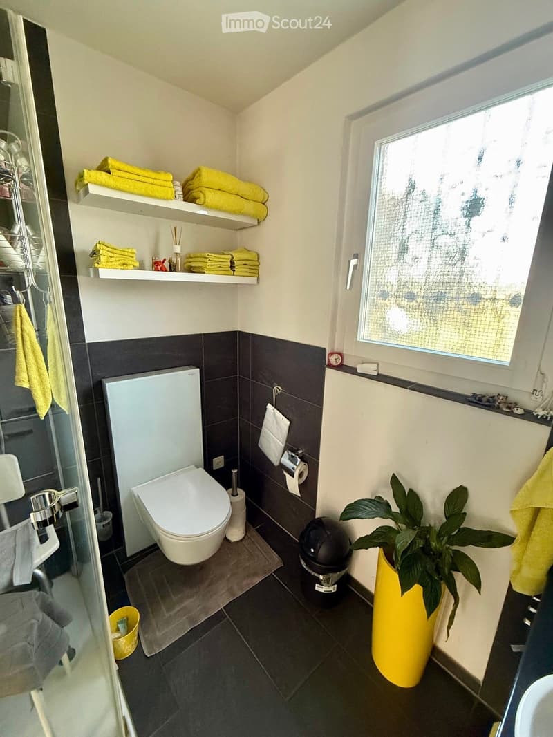 Bathroom with white toilet, shower, shelves with towels, window, trash can, plant, and tiled floor.
