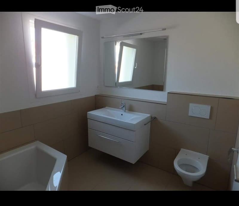 Modern bathroom with a white sink, mirror, toilet, and bathtub. Light-colored tiles and a window provide natural light.