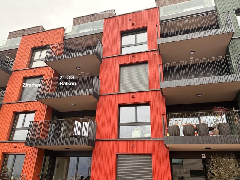 Modern multi-story building with red wooden facade, multiple balconies, large windows, and modern railings.