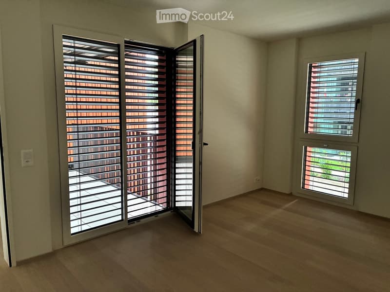 Empty room with light-colored walls, wooden flooring, and large windows with blinds. The room has a sliding glass door leading to a balcony or outdoor area.