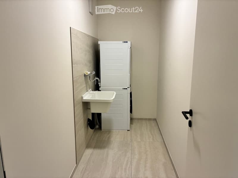 Small bathroom with a white sink, faucet, toilet paper holder, and a white washing machine. Light-colored tiled walls and floor.