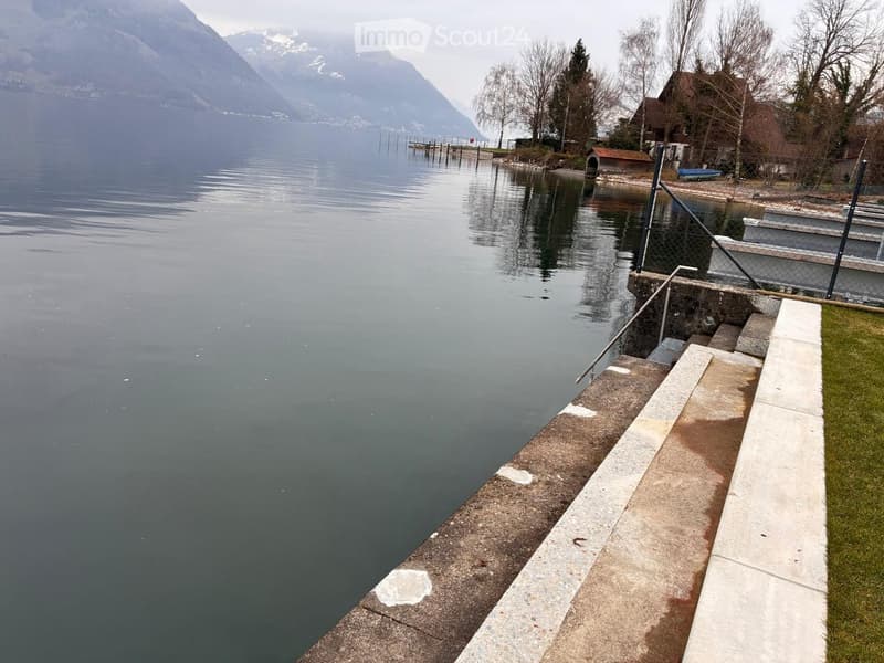 A calm lake with a concrete dock extending into the water, surrounded by grassy areas and distant mountains.