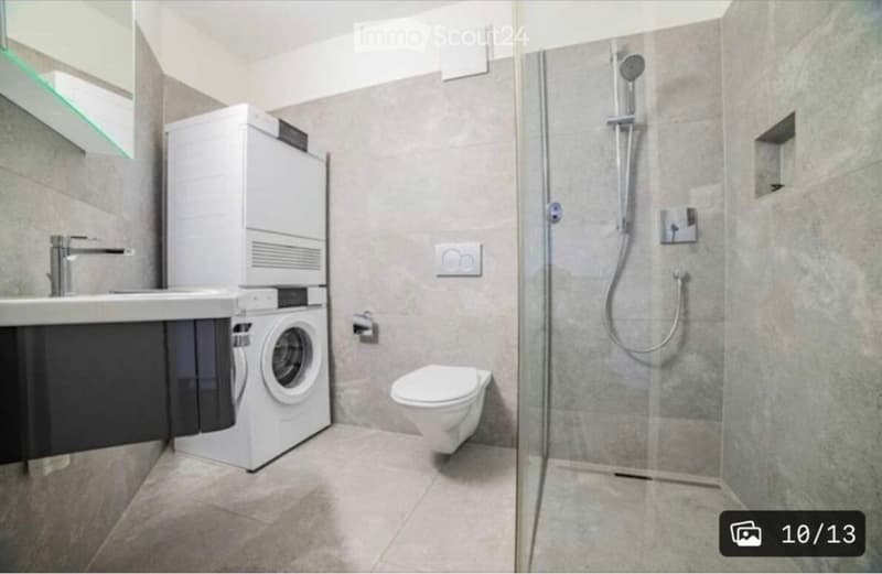 Modern bathroom with a white washing machine, sink, toilet, and glass-enclosed shower. The walls are tiled in a neutral gray color, and the floor is covered with light gray tiles.