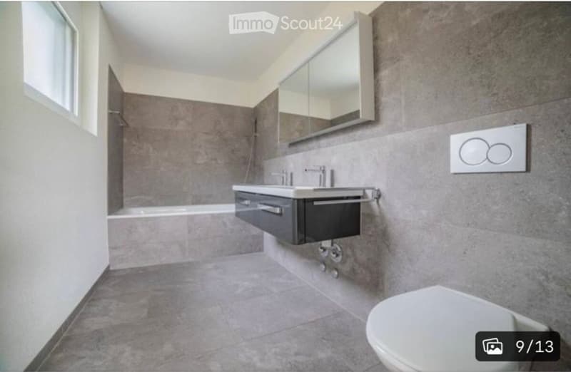Modern bathroom with grey tiled walls and floor, a white toilet, a sink with a dark cabinet, and a large mirror above the sink.