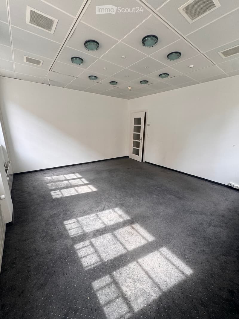 Empty room with white walls, grey carpeted floor, and a white door with glass panels. The ceiling features several round light fixtures and air vents.