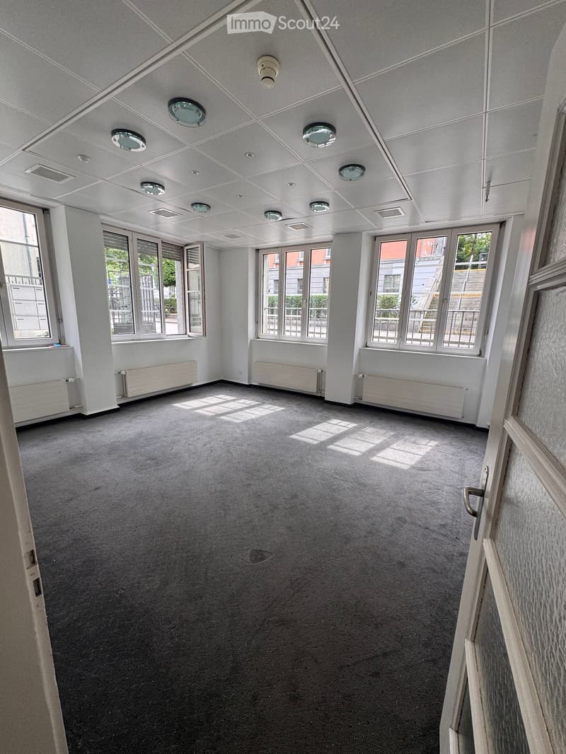 Empty room with grey carpet, white walls, large windows, radiators, and recessed ceiling lights.