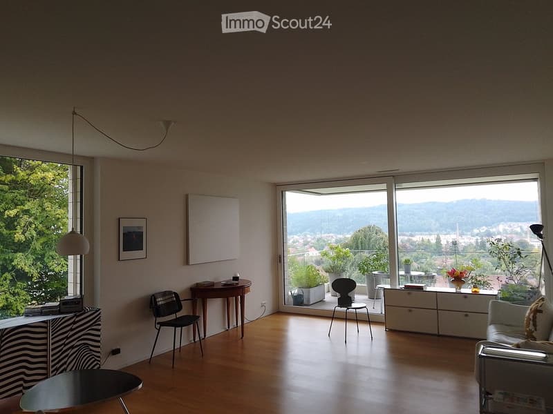 Bright living room with wooden floor, large windows offering a scenic view, sofa, chairs, tables, and decorative items.