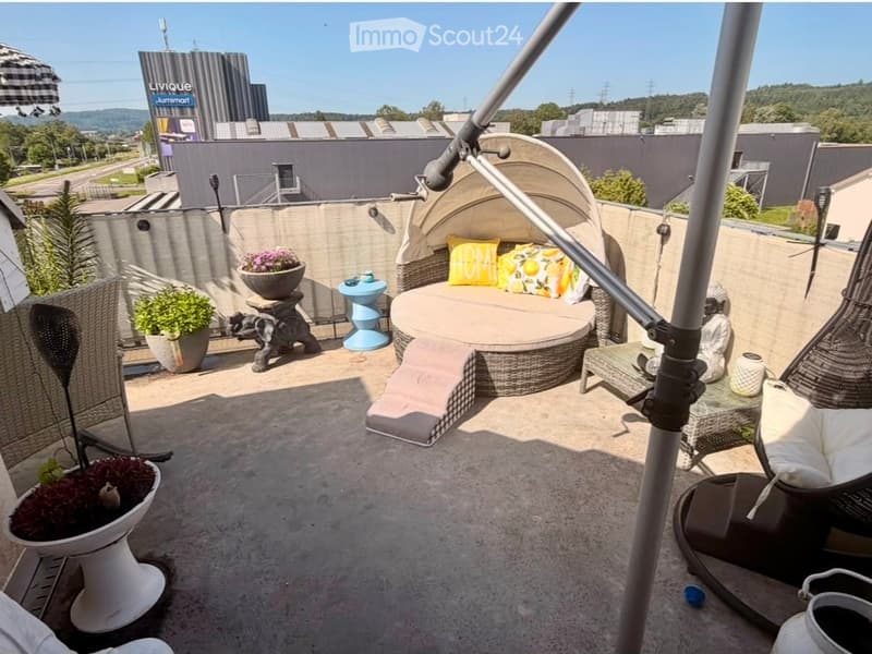 A modern rooftop terrace with a round wicker sofa, colorful cushions, a small table, potted plants, and an umbrella.