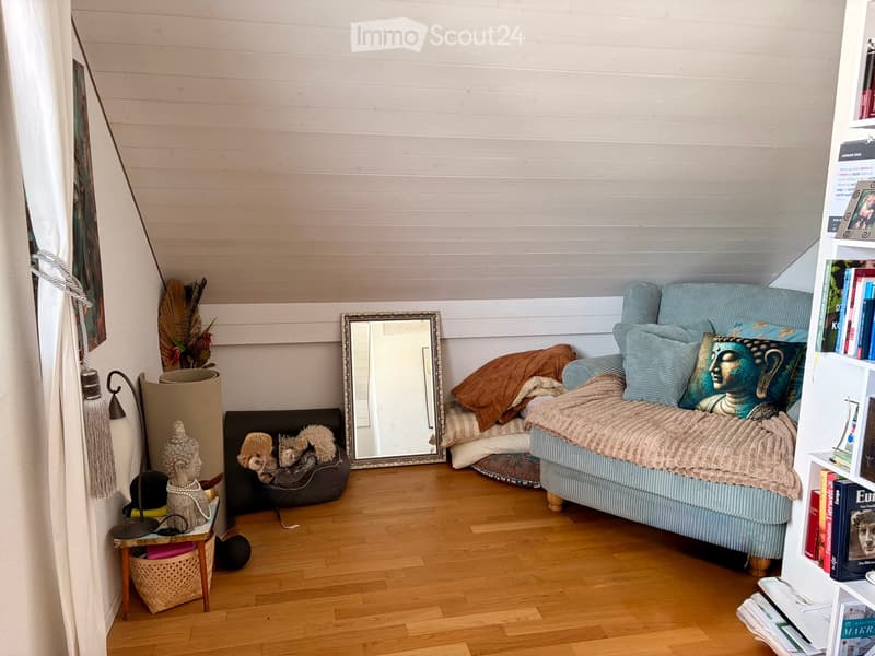 Cozy bedroom with a light blue sofa, wooden floor, mirror, small table with a lamp, bookshelf, decorative statue, and a pet bed.