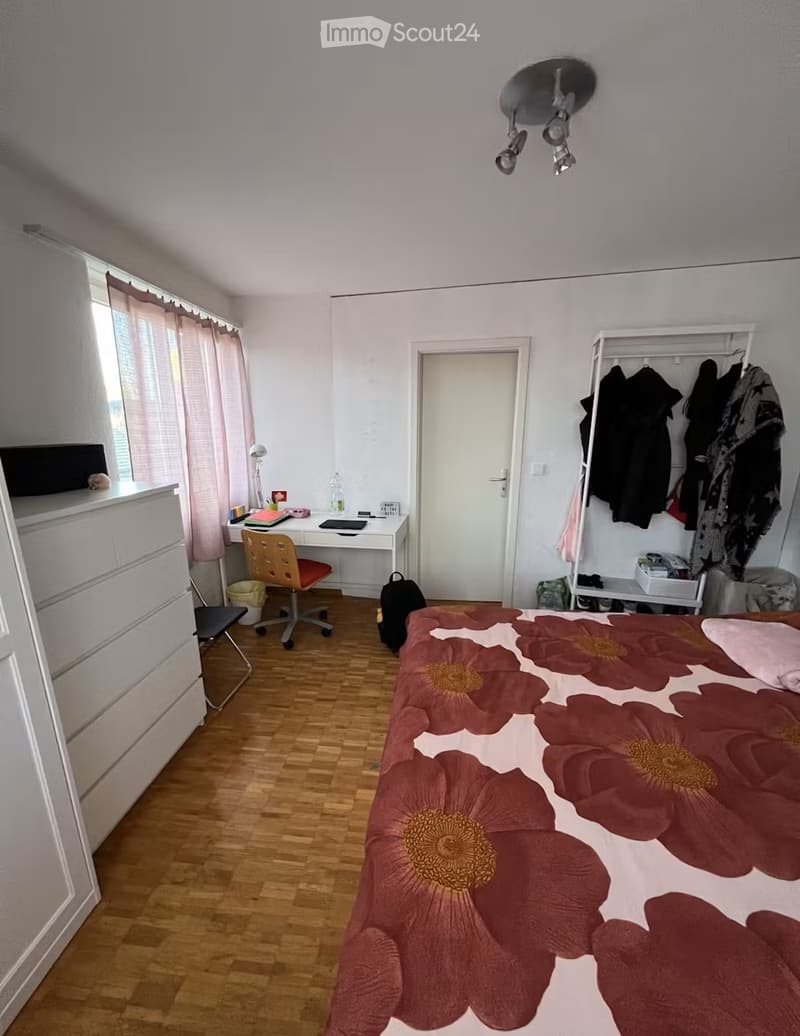 A well-lit bedroom with wooden flooring, a large bed with a floral duvet, a desk with a chair, a wardrobe with hanging clothes, and a window with curtains.