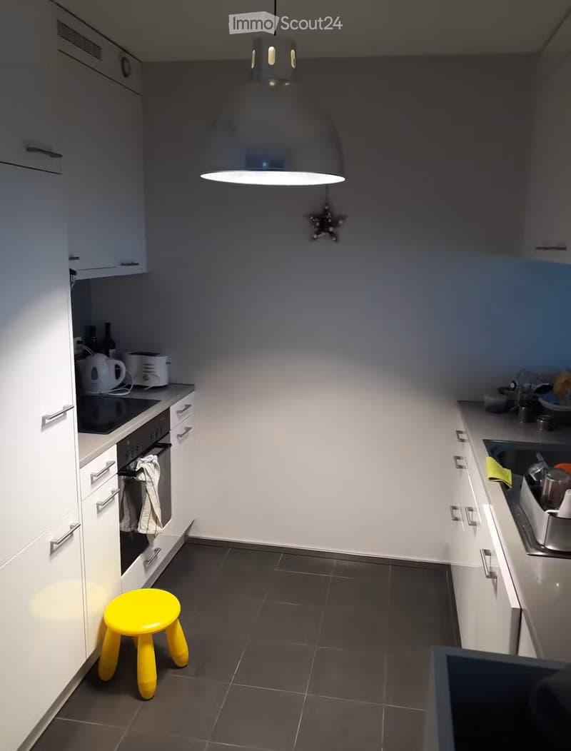 Modern kitchen with white cabinets, dark tiled floor, and a yellow stool in the center. Features an oven, sink, and various kitchen appliances on the counters.