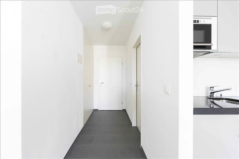 A narrow, white-walled hallway with a dark tiled floor, leading to a closed door. A small kitchen area is visible on the right, featuring white cabinets, a microwave, and a sink.