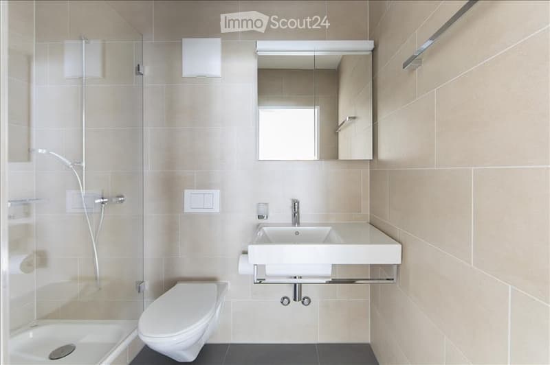 Modern bathroom with beige tiled walls, a white toilet, a sink with a mirror above it, and a glass-enclosed shower area.