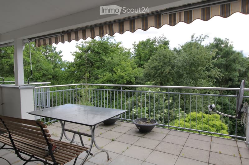 Balcony with a table, bench, and potted plants, overlooking a green, tree-filled landscape.
