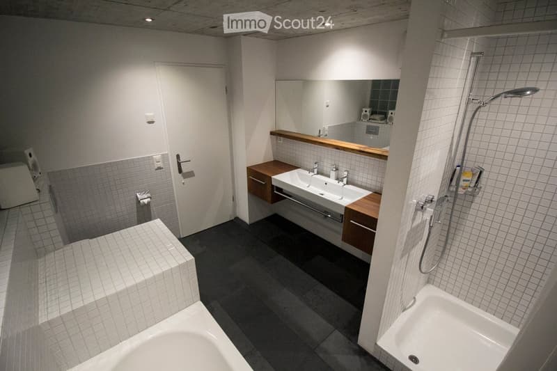 Modern bathroom with a double sink, bathtub, shower, and built-in wooden cabinetry. The space features tiled walls and a dark floor, with a large mirror above the sink.