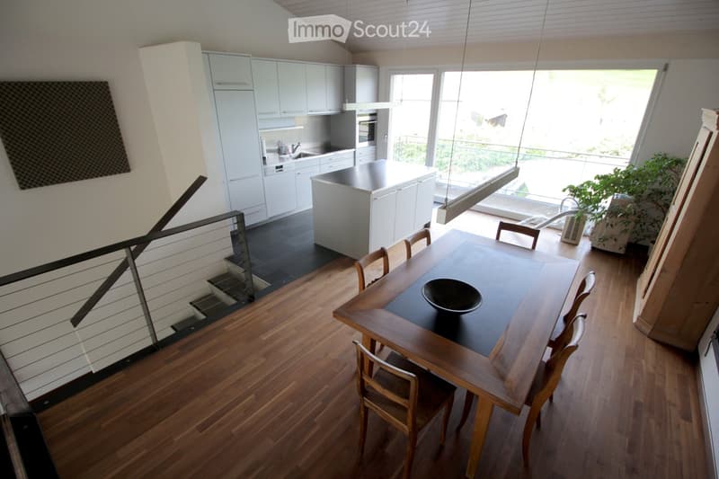 Open-plan kitchen and dining area with light-colored cabinets, a central island, and a wooden dining table with chairs. The space features wooden flooring and large windows with a view of a balcony.