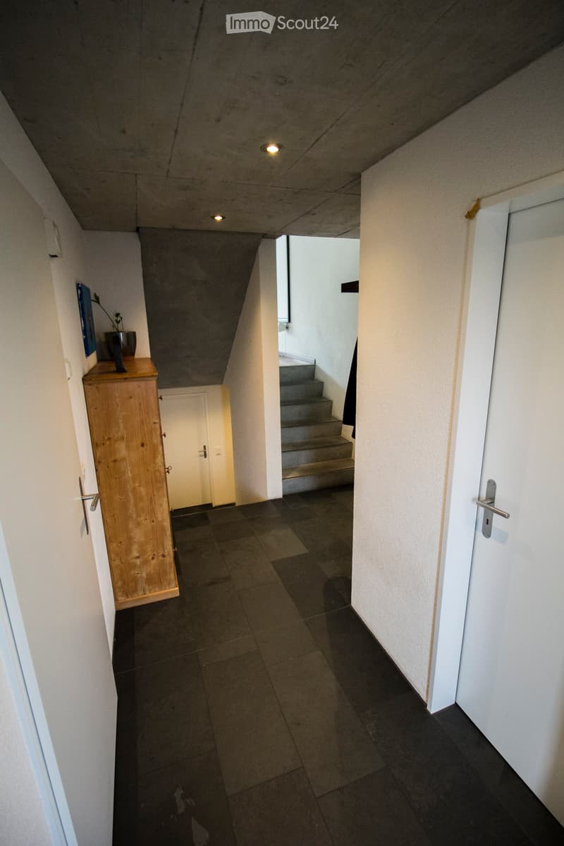 Modern hallway with dark tiled floor, white walls, recessed ceiling lights, and a small wooden cabinet with a plant on top. A staircase leads upwards, and a door is visible on the right.