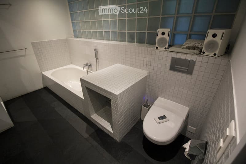 Modern bathroom with a bathtub, toilet, sink, and speakers on the counter. The walls are tiled, and there is a remote control on the toilet seat.