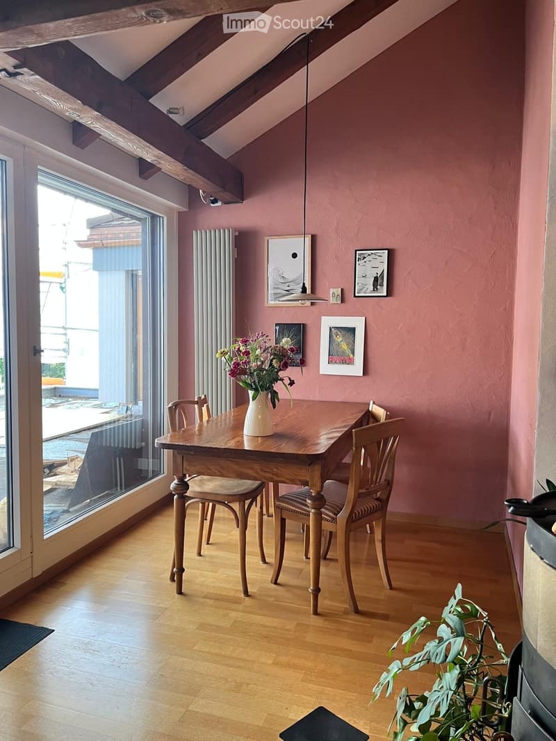 Dining area with wooden table, chairs, vase with flowers, and artwork on walls. Large window with view.