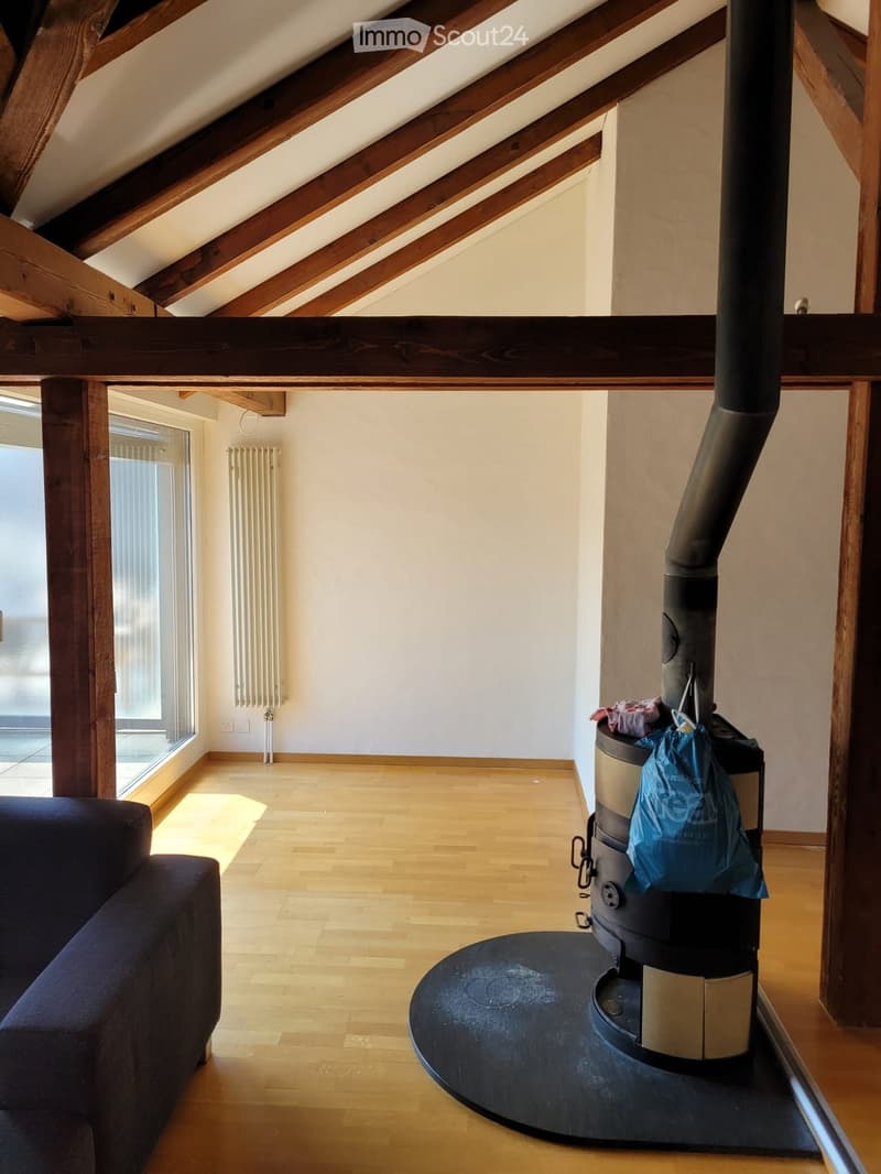 Living room with wooden floor, exposed wooden beams, wood stove, sofa, radiator, and large window with blinds.