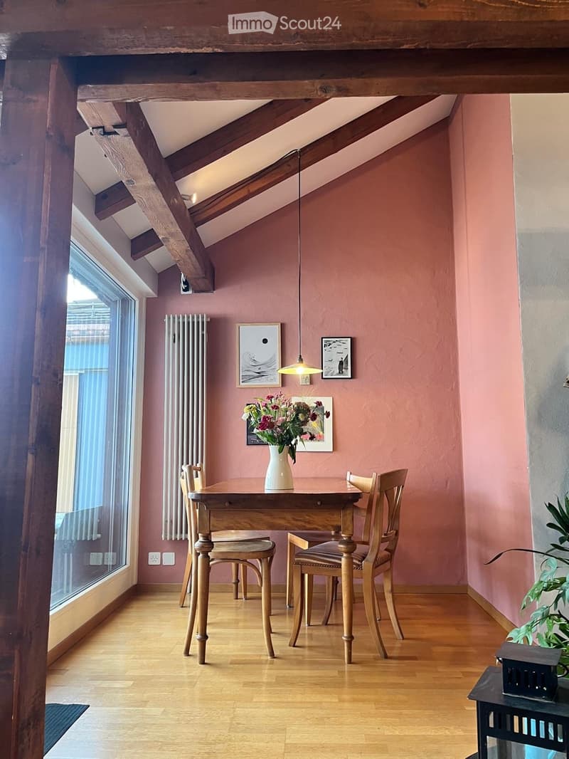 Cozy attic dining area with wooden beams, pink walls, and large window. Includes a wooden dining table, four chairs, a vase with flowers, and artwork on the walls.