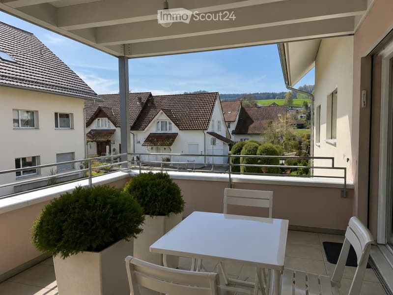 Balcony with a white table and chairs, surrounded by greenery and a scenic view of neighboring houses and countryside.