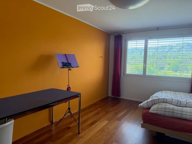 Bright bedroom with wooden flooring, orange accent wall, large window with view, bed, table, and music stand.