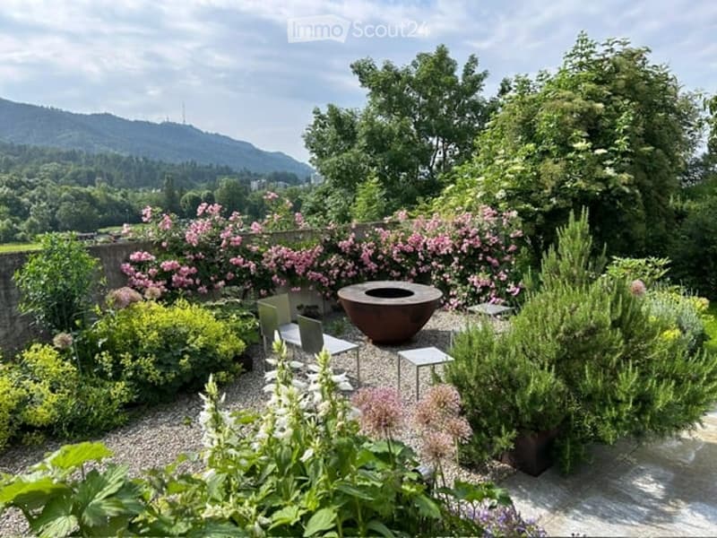 Well-maintained garden with a variety of flowering plants, shrubs, and trees. Features include a stone wall, a large decorative bowl, and a small seating area with chairs and a table.