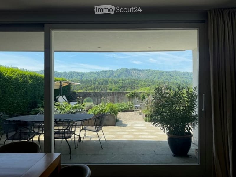 Balcony with large glass doors opening to a scenic outdoor area, featuring a table and chairs, potted plants, and a view of a landscaped garden and distant hills.