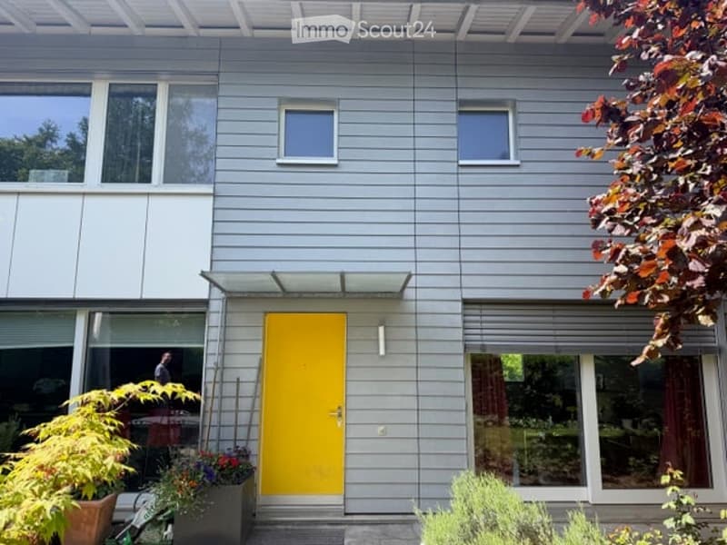 Modern two-story house with grey siding, large windows, and a bright yellow front door. The entrance is flanked by potted plants and small shrubs.