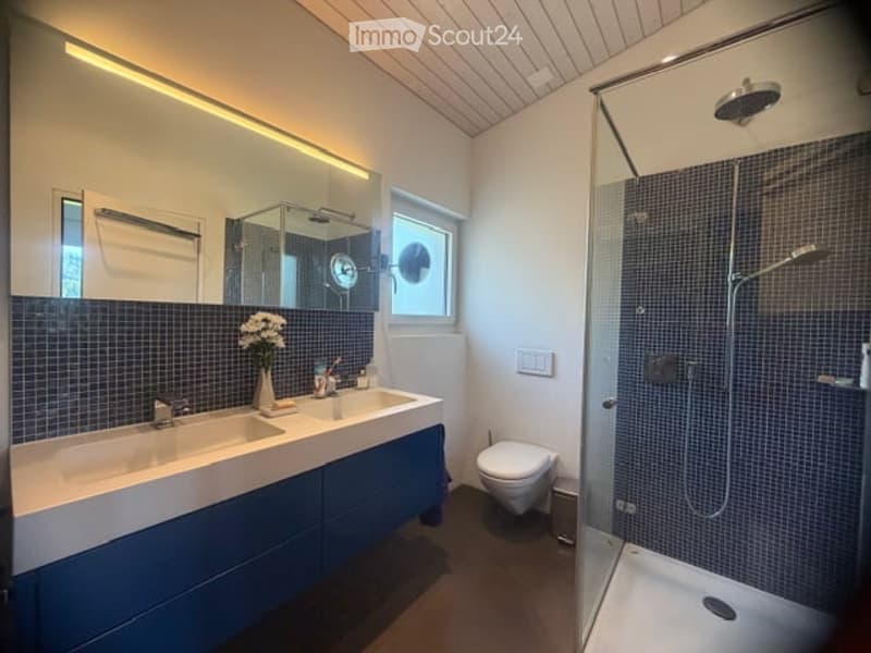 Modern bathroom with double sink, large mirror, shower, and toilet. Blue tiles on lower half of walls, white upper half, and wooden ceiling.