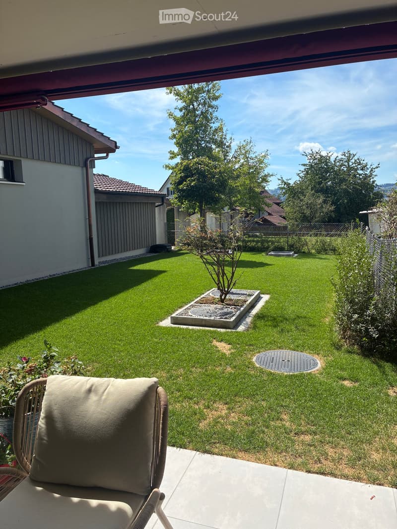 Well-maintained garden with a lawn, small tree, and a covered patio area with a chair and cushion.