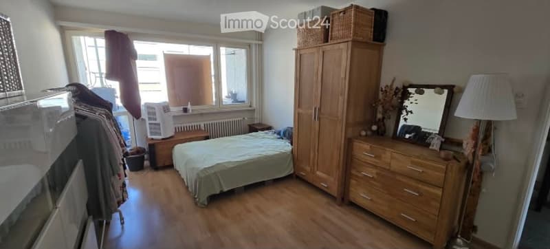 Cozy bedroom with wooden flooring, a single bed with a green duvet, a large wooden wardrobe, a dresser with a mirror, a floor lamp, and a wicker basket on top of the wardrobe. A window with blinds and an air conditioner unit is visible.