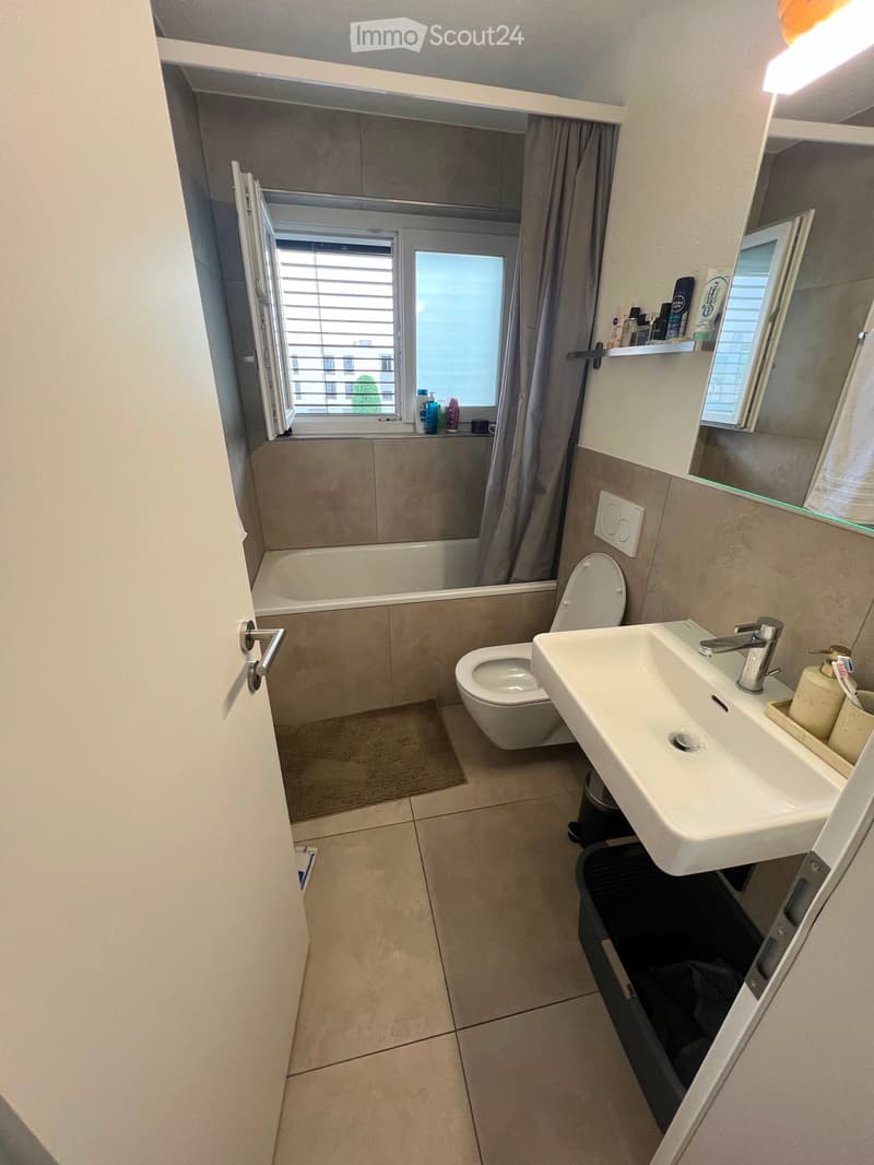 Modern bathroom with a white sink, toilet, bathtub, and mirror. The room features tiled walls and floor, a window with blinds, and a small shelf with toiletries.