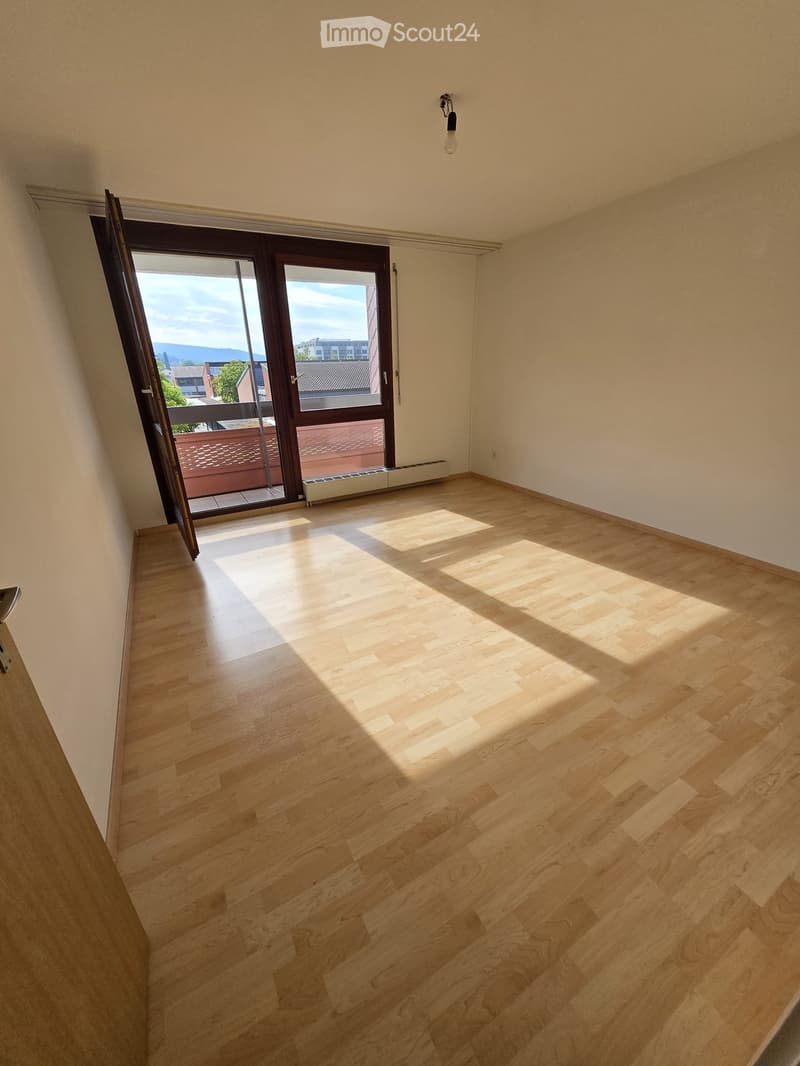 Empty room with wooden flooring, large sliding glass door leading to a balcony, and natural light streaming in.