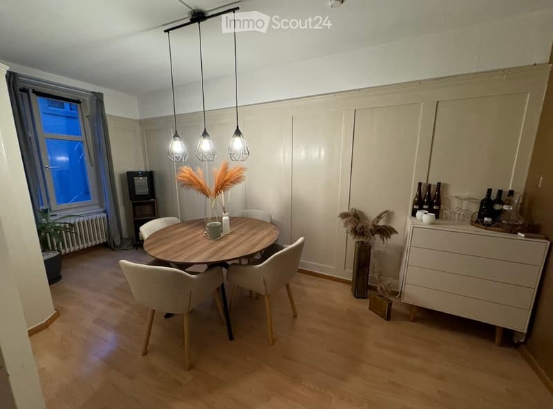 Dining area with a round wooden table, four white chairs, pendant lights, and decorative items including vases and bottles on a sideboard.