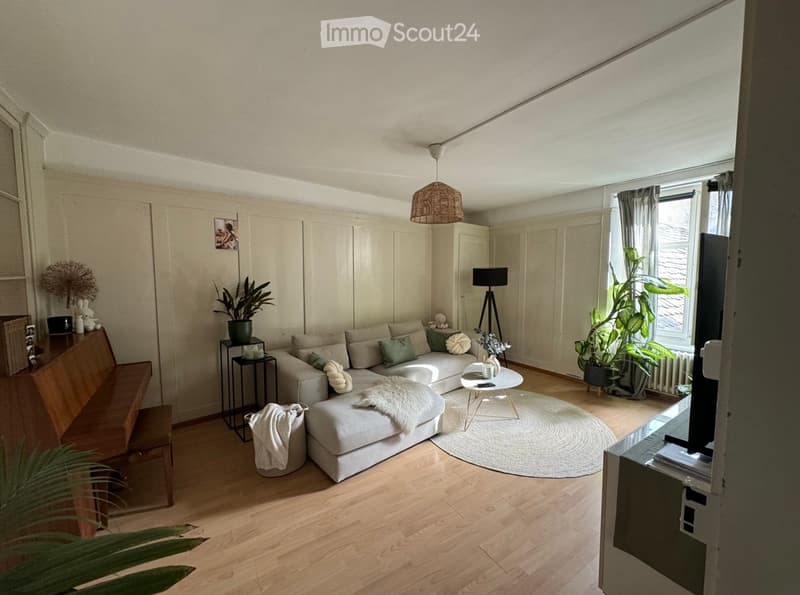 Modern living room with light-colored sofa, coffee table, bench, floor lamp, and plants. The room features wooden flooring and large windows with curtains.